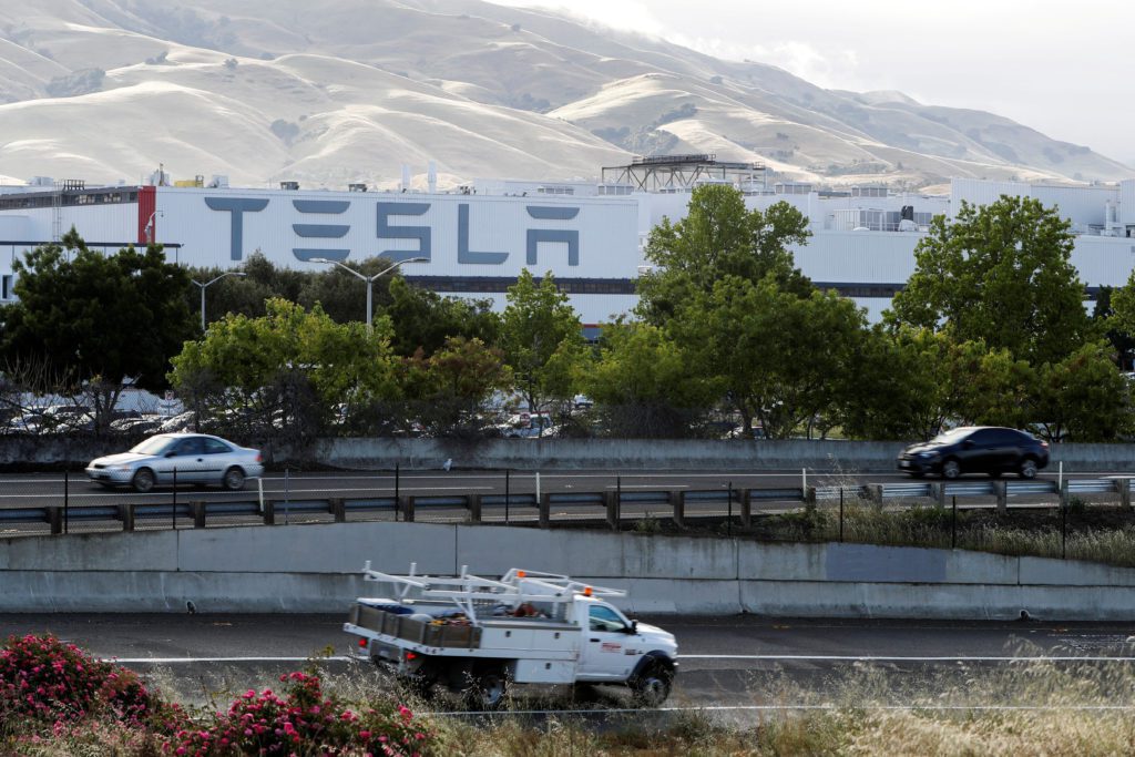 Fábrica Tesla com logo visível, carros em movimento em rodovia abaixo, e montanhas ao fundo.