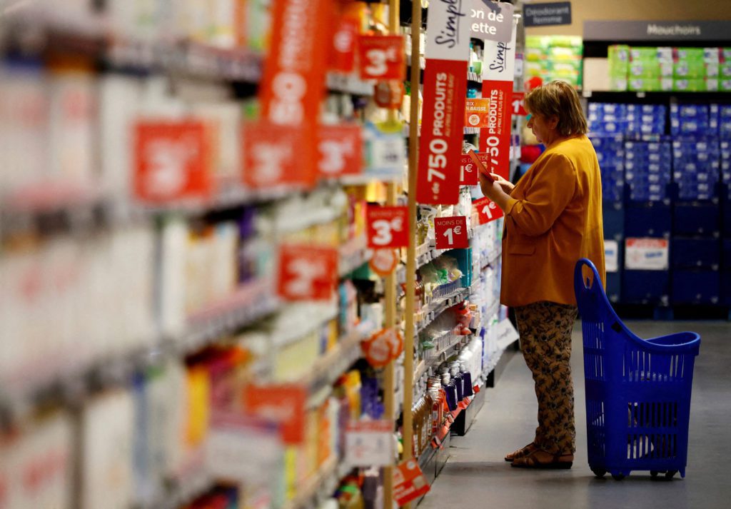 Mulher com carrinho de compras azul examina produtos em prateleira de supermercado.