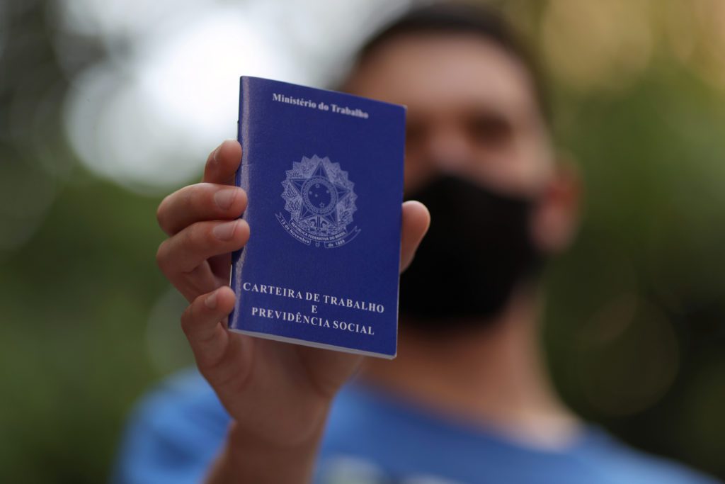 Mão segura Carteira de Trabalho azul. Pessoa com máscara desfocada ao fundo.