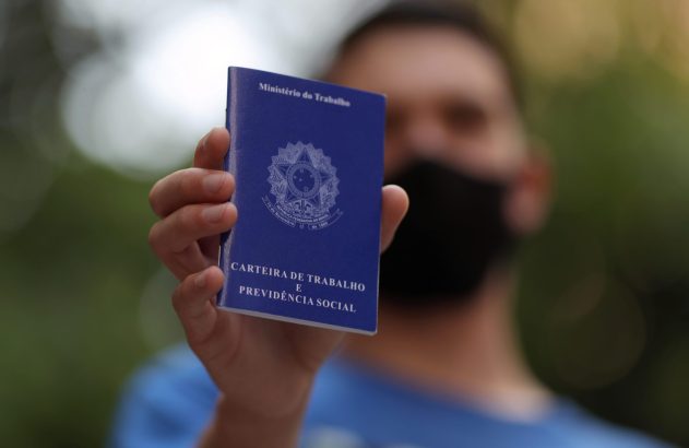 Mão segura Carteira de Trabalho azul. Pessoa com máscara desfocada ao fundo.