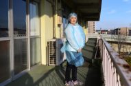 Mulher com touca, avental e luvas de proteção azul em um corredor externo ensolarado.