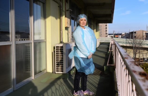 Mulher com touca, avental e luvas de proteção azul em um corredor externo ensolarado.