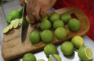 Mãos cortando limões verdes em uma tábua de madeira com uma faca, ao lado de um espremedor.
