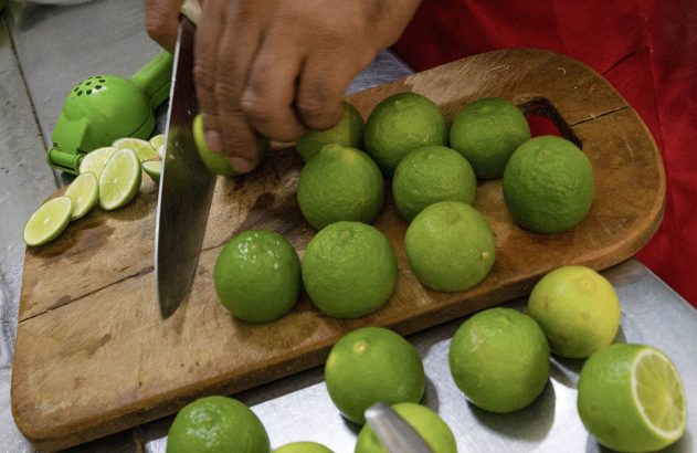 Mãos cortando limões verdes em uma tábua de madeira com uma faca, ao lado de um espremedor.