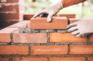 Mãos com luvas assentando um tijolo em uma parede de tijolos com argamassa.