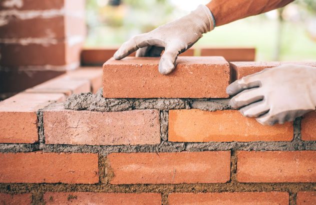 Mãos com luvas assentando um tijolo em uma parede de tijolos com argamassa.
