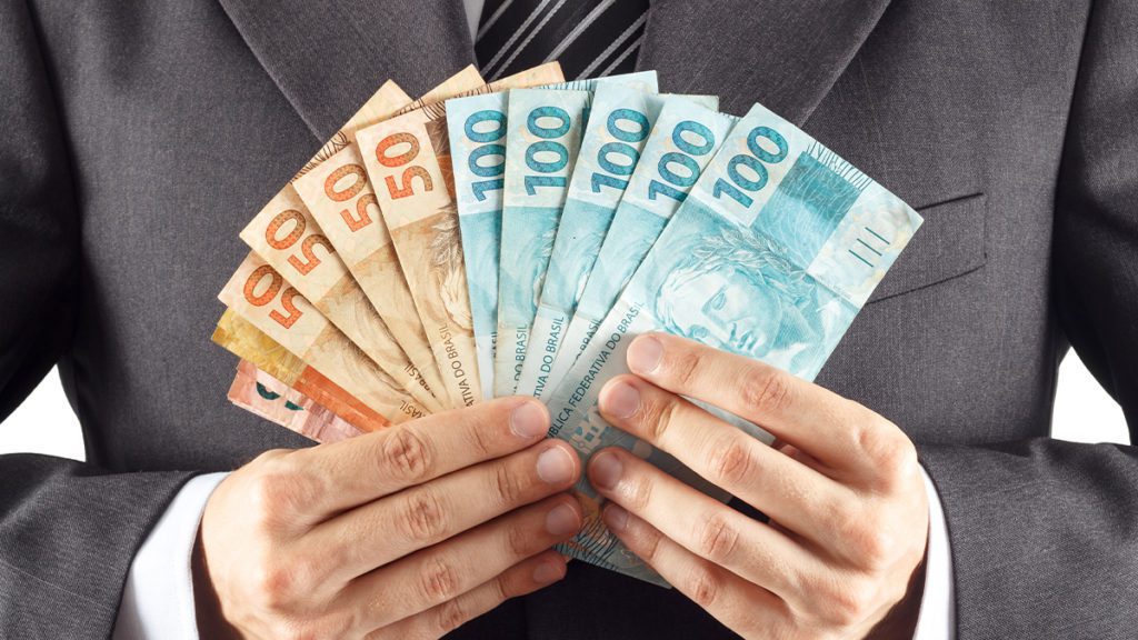Pessoa de terno segura um leque de notas de Real, incluindo notas de 50 e 100.