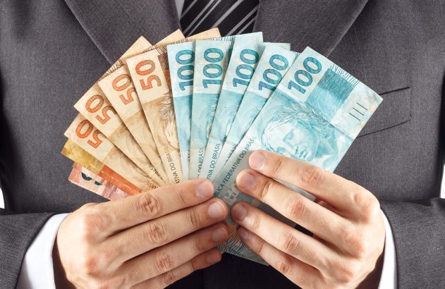 Pessoa de terno segura um leque de notas de Real, incluindo notas de 50 e 100.