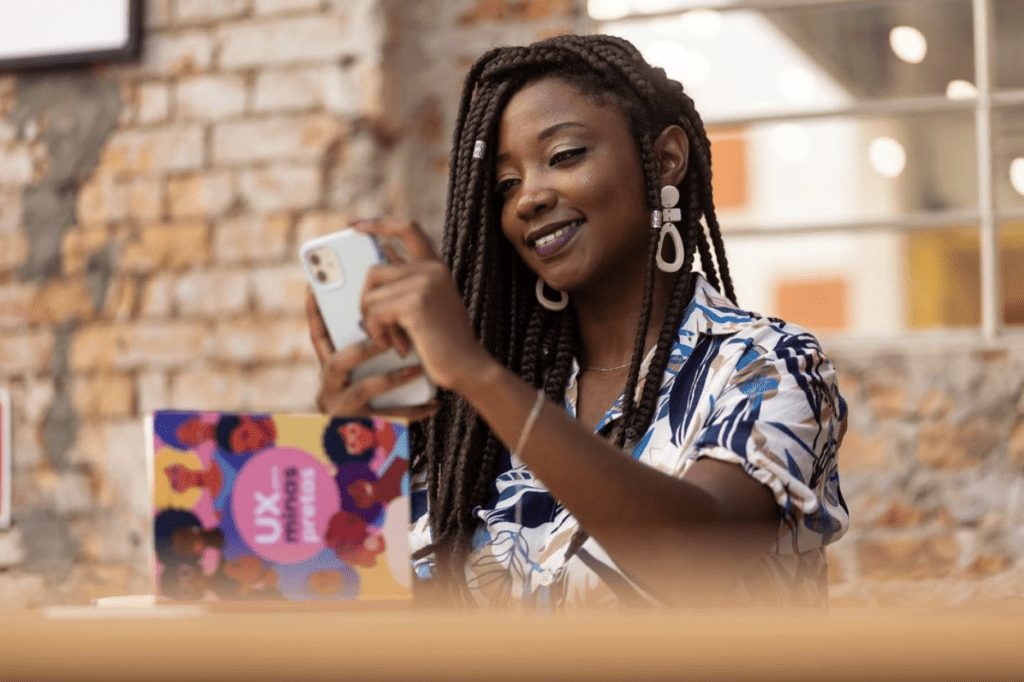 Mulher negra de tranças sorri usando um smartphone. Na mesa, uma caixa colorida com "UX minas pretas".
