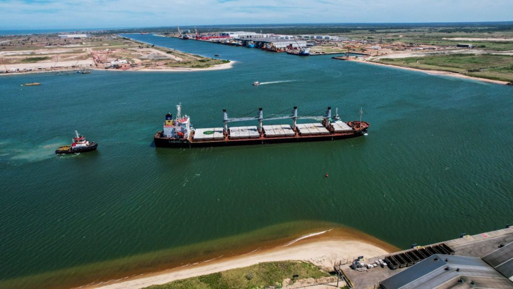 Navio cargueiro em canal de porto, assistido por rebocador. Complexo industrial ao fundo.