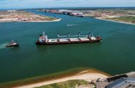 Navio cargueiro em canal de porto, assistido por rebocador. Complexo industrial ao fundo.