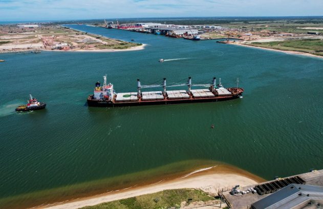 Navio cargueiro em canal de porto, assistido por rebocador. Complexo industrial ao fundo.
