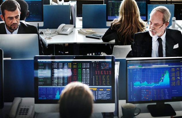 Profissionais em ambiente de trabalho com monitores exibindo informações de mercado.