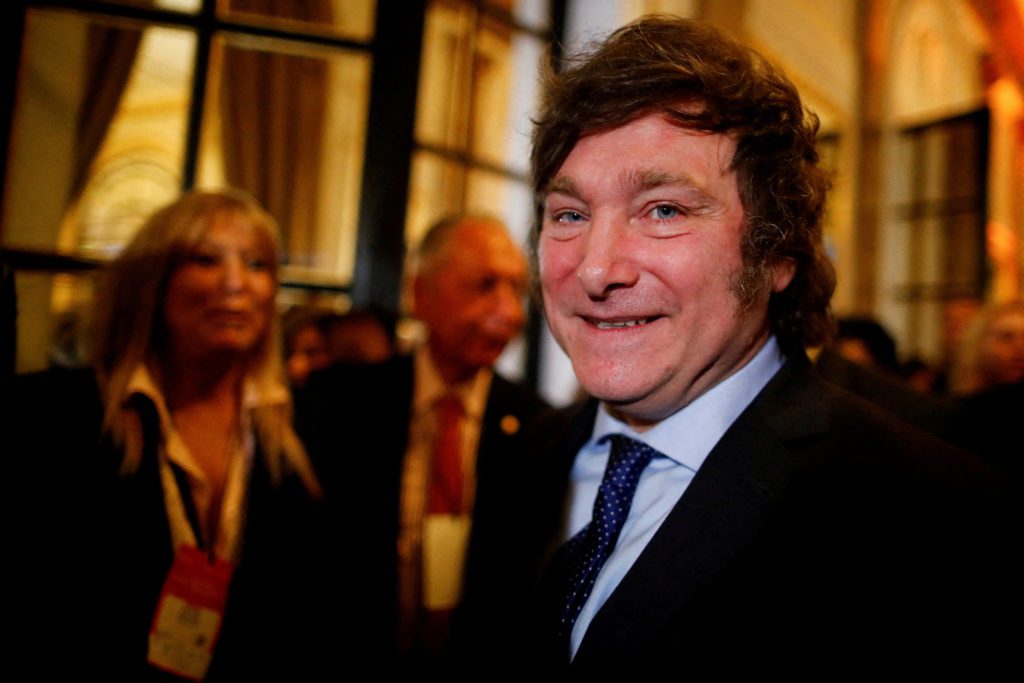 Javier Milei sorri para a câmera, vestindo terno e gravata, em evento com fundo desfocado de pessoas.