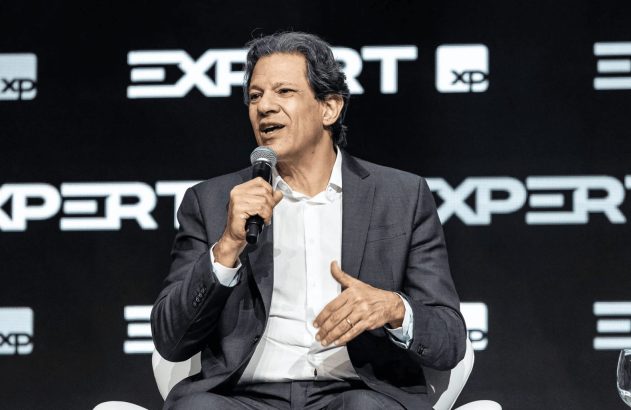 Palestrante de terno e camisa branca fala ao microfone em evento XP EXPERT.
