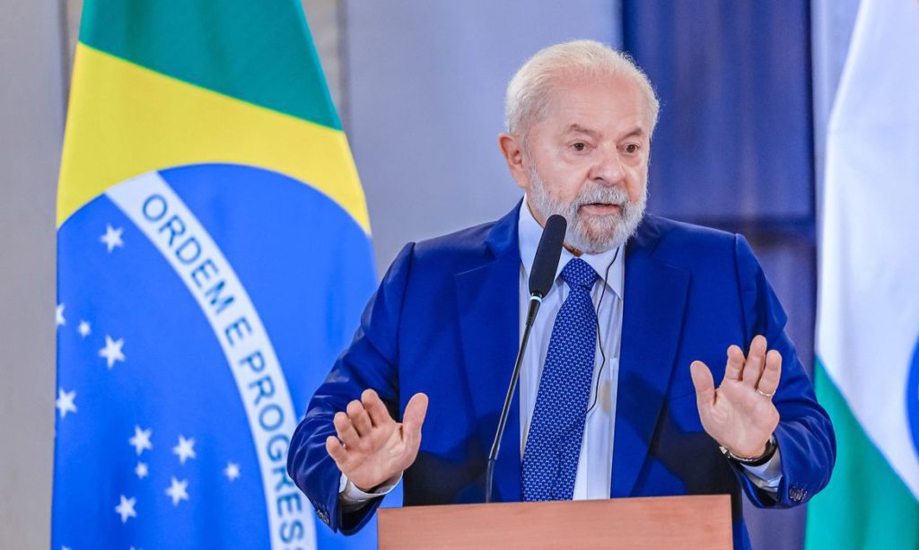 Homem de terno azul com barba branca fala em pódio, gesticulando. Bandeira do Brasil ao fundo.