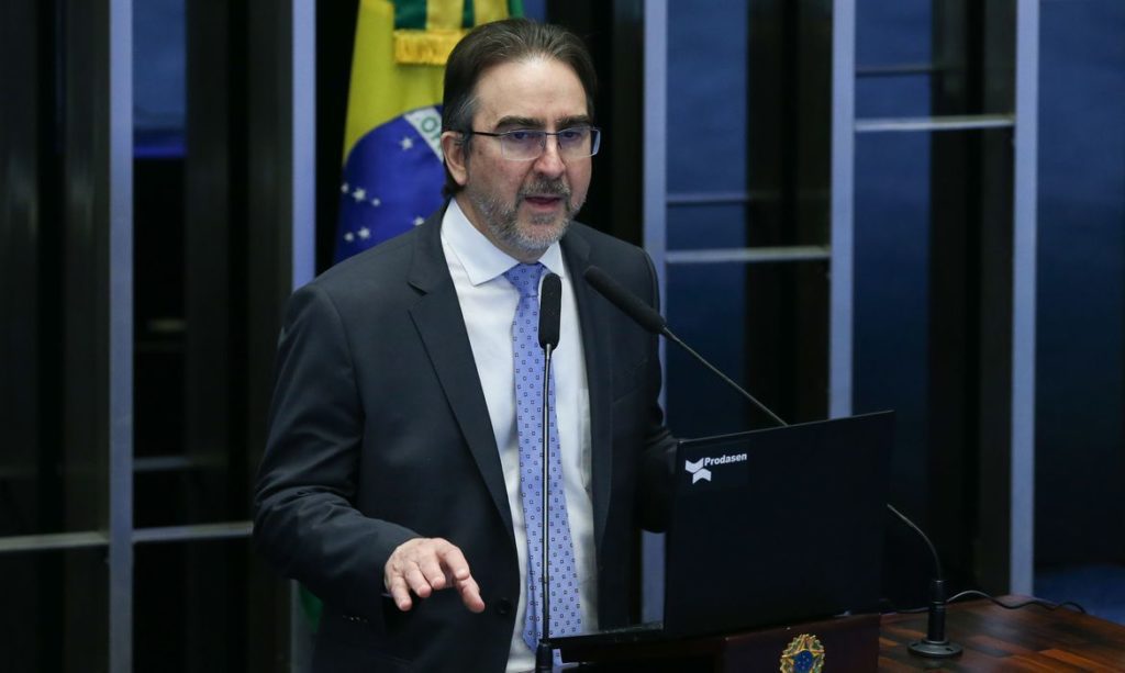 Homem de terno e gravata azul fala ao microfone em evento. Bandeira do Brasil e laptop Prodasen no fundo.