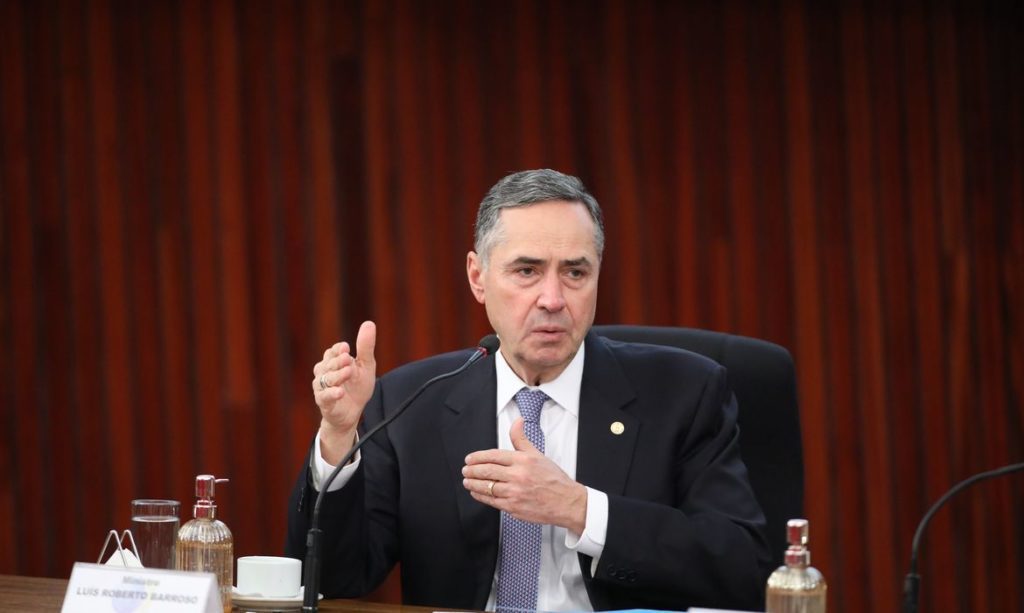 Ministro Luís Roberto Barroso, de terno, gesticula e fala sentado à mesa.