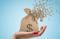 Mão segura saco de dinheiro com cifrão, se desfazendo com notas de dólar voando. Fundo azul.