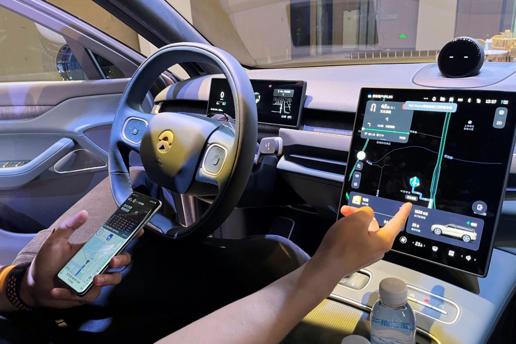 Mãos interagem com painel de carro moderno. Uma toca a tela touchscreen com mapa, outra segura celular.