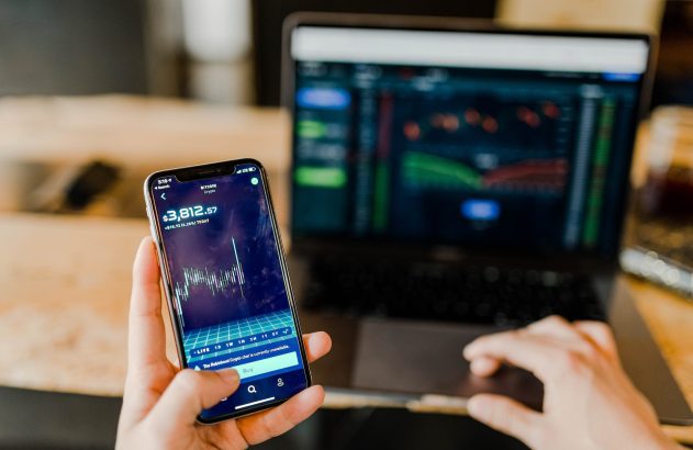 celular com gráfico de ações em uma mão, com um notebook ao fundo. A imagem ilustra o termo dividend yield