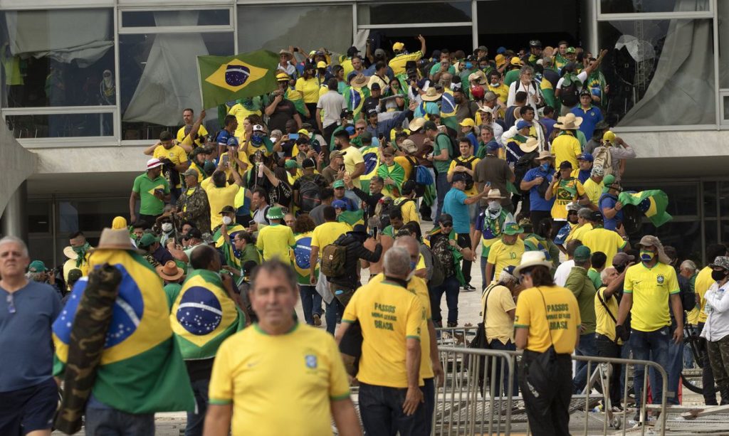 Multidão com bandeiras do Brasil e camisas amarelas em prédio com vidro quebrado.