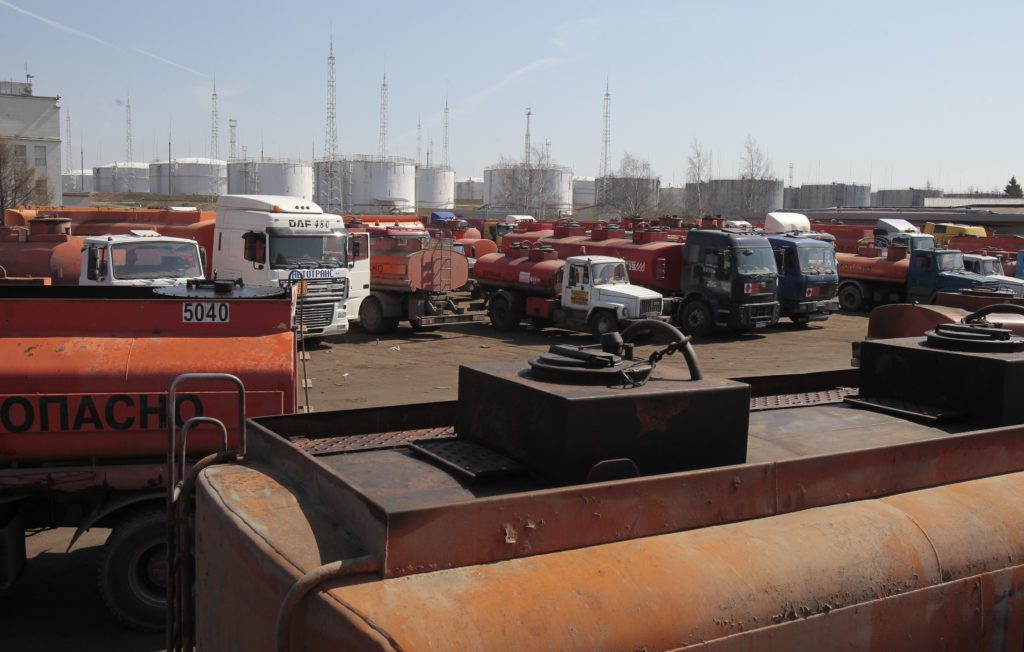Muitos caminhões-tanque parados em pátio de combustível com tanques de armazenamento industriais ao fundo.