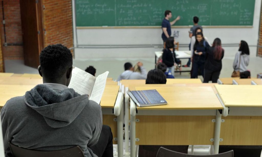 Estudante lendo em sala de aula, com professor e colegas no fundo perto do quadro.