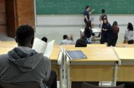 Estudante lendo em sala de aula, com professor e colegas no fundo perto do quadro.