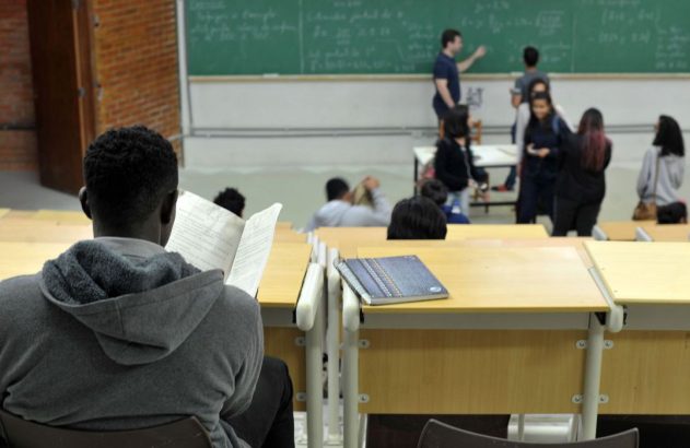 Estudante lendo em sala de aula, com professor e colegas no fundo perto do quadro.