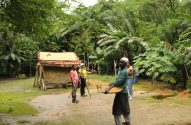 Grupo de pessoas em floresta tropical exuberante com cabana rústica. Um homem à frente segura uma prancheta.