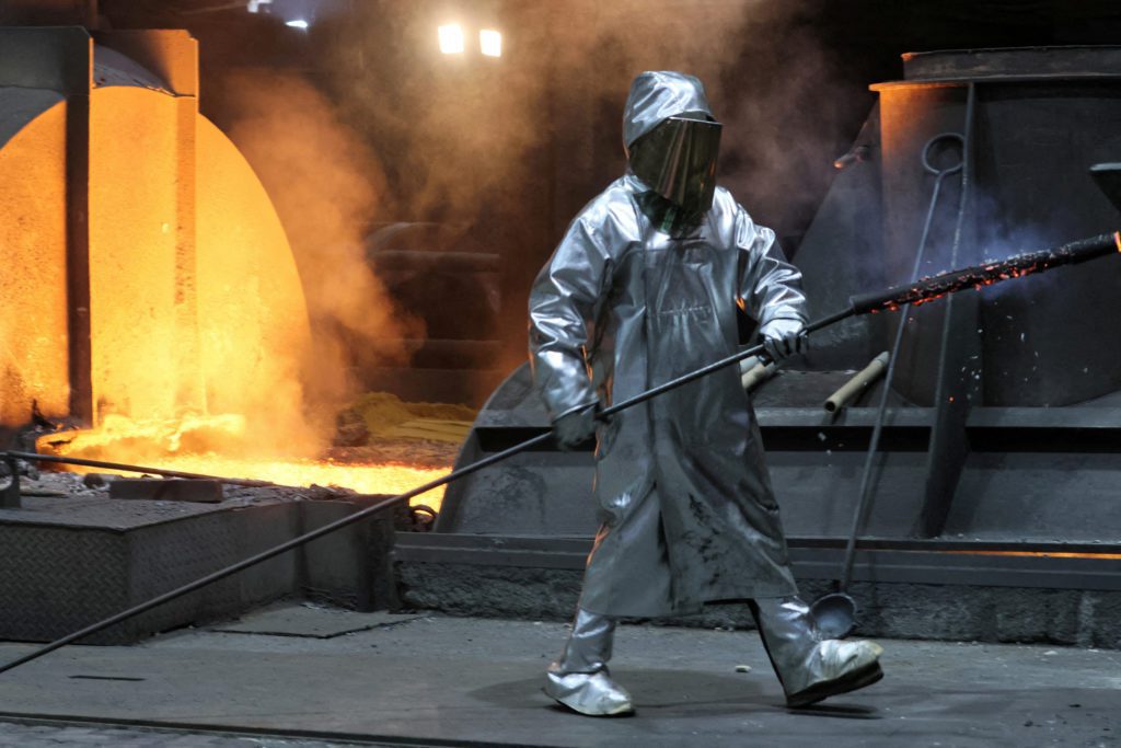 Trabalhador de forno em traje protetor prateado, manuseando uma ferramenta longa perto de metal líquido incandescente.