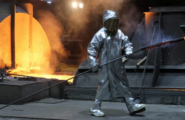 Trabalhador de forno em traje protetor prateado, manuseando uma ferramenta longa perto de metal líquido incandescente.