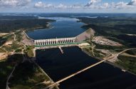 Vista aérea: Imensa usina hidrelétrica em operação, com barragem, vasto reservatório e floresta densa ao redor.