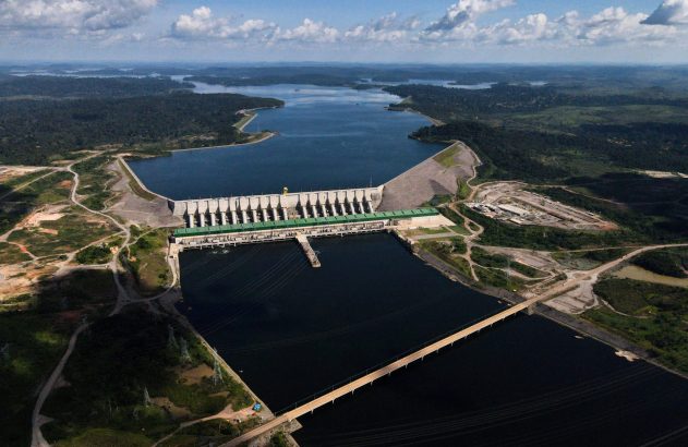 Vista aérea: Imensa usina hidrelétrica em operação, com barragem, vasto reservatório e floresta densa ao redor.