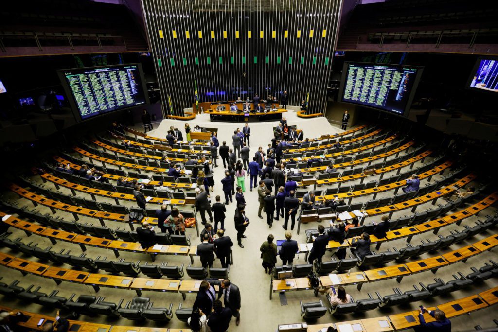 Amplo plenário brasileiro: parlamentares conversam e circulam entre as cadeiras. Telas exibem listas e dados.