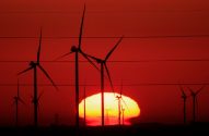 Silhuetas de turbinas eólicas contra um pôr do sol vermelho intenso com o sol amarelo no horizonte e linhas de energia.