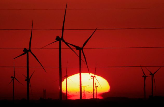Silhuetas de turbinas eólicas contra um pôr do sol vermelho intenso com o sol amarelo no horizonte e linhas de energia.