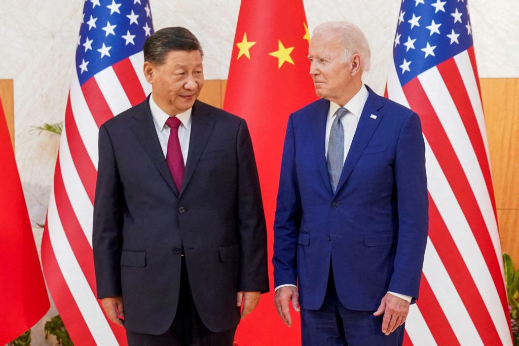 Presidentes Xi Jinping e Joe Biden se olham, ladeados por bandeiras dos EUA e China.