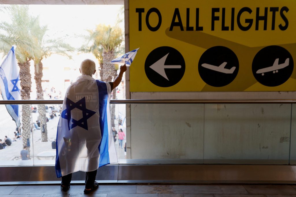 Pessoa de costas com bandeira de Israel como capa, segurando pequena bandeira. Em aeroporto, com sinal "TO ALL FLIGHTS".