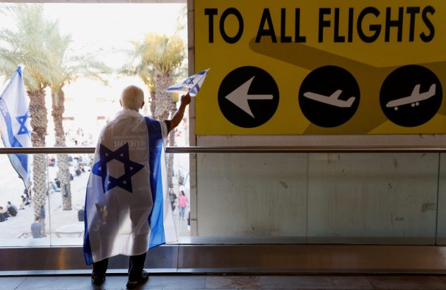 Pessoa de costas com bandeira de Israel como capa, segurando pequena bandeira. Em aeroporto, com sinal "TO ALL FLIGHTS".