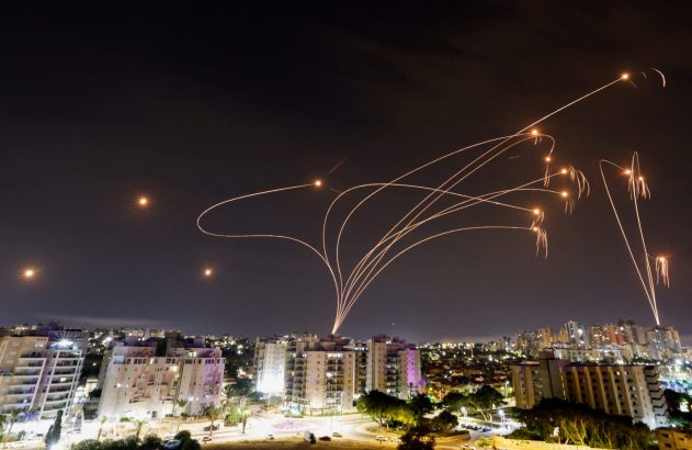 Céu noturno sobre cidade com múltiplas trilhas de luz de foguetes e pontos de interceptação brilhantes.