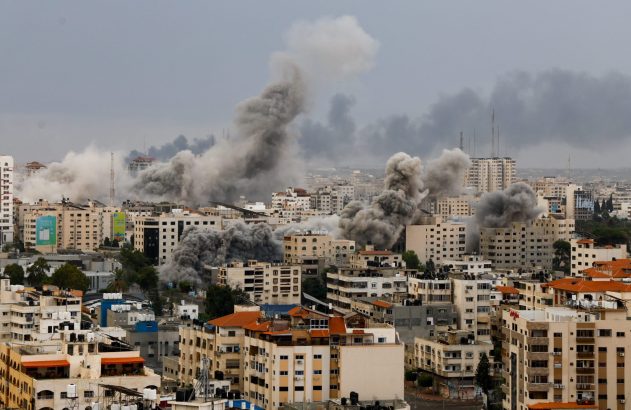 Cenário urbano com fumaça cinza e densa de múltiplas explosões subindo entre os edifícios.