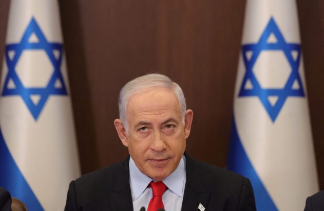 Um homem de terno e gravata vermelha, com duas bandeiras de Israel ao fundo, olhando para a frente.