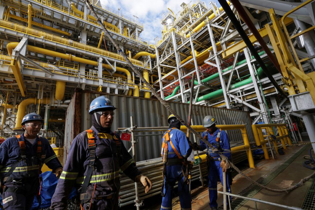 Trabalhadores com EPIs em plataforma industrial com tubulações, manuseando cabos.