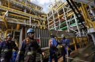Trabalhadores com EPIs em plataforma industrial com tubulações, manuseando cabos.