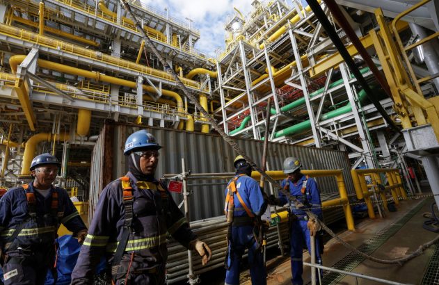 Trabalhadores com EPIs em plataforma industrial com tubulações, manuseando cabos.