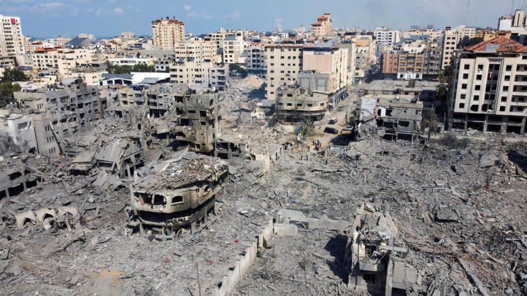 Vista de cima de uma área urbana devastada, coberta por escombros e ruínas de edifícios destruídos, com alguns prédios de pé ao fundo.