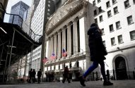 Pessoas caminhando em Wall Street, em frente ao icônico prédio da Bolsa de Valores de NY com colunas e bandeiras dos EUA.