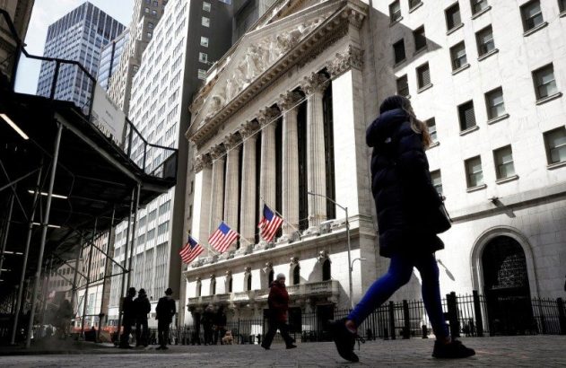 Pessoas caminhando em Wall Street, em frente ao icônico prédio da Bolsa de Valores de NY com colunas e bandeiras dos EUA.